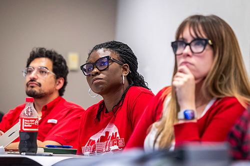 Three students, one male on the left and two females with red shirts looking intently in a classroom
