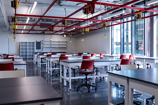 Several groups of tables in a teaching labratory with electrical plugs hanging from red overhead frames