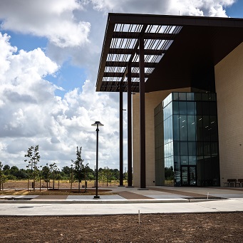 The glass entrance of a three story brick building. A brown metal awning is over the entrance, and clouds can be seen in the blue sky in the background