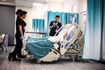 Three nursing students stand around a hospital bed with a high-fidelity mannequin.