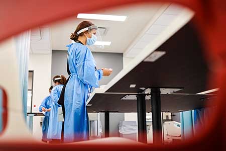 A groupd of nursing students wearing surgical gowns work at tables.