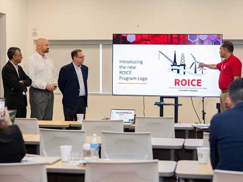 Four men stand in from of a classroom. One with a red shirt points to a presentation slide that states "Introducting the new ROICE Program Logo".