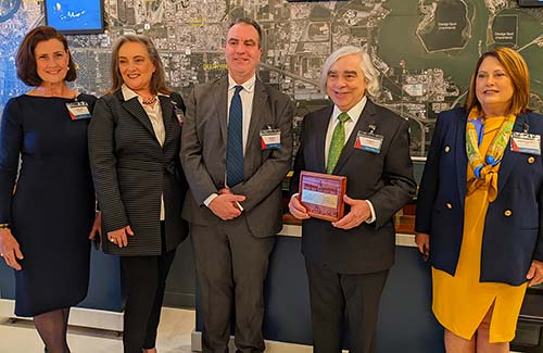 Five people in business attire stand in front of large map. The second person from the right holds a plaque.