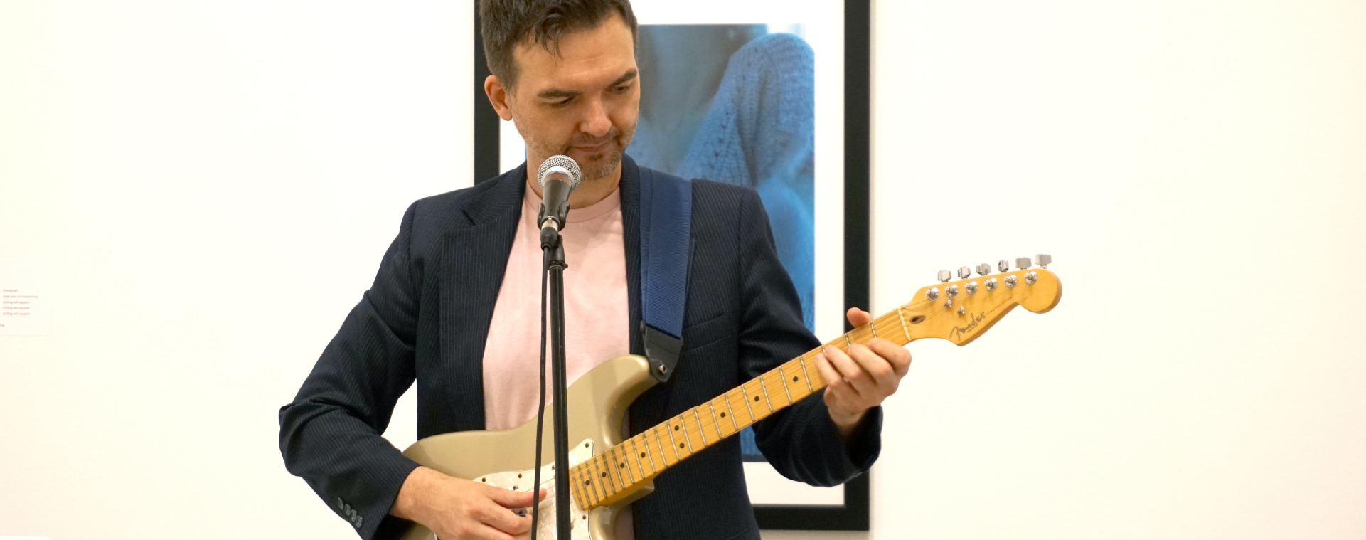 Close up of a performer with cropped dark hair, wearing a dark blazer and pale t-shirt behind a microphone playing an electric guitar.