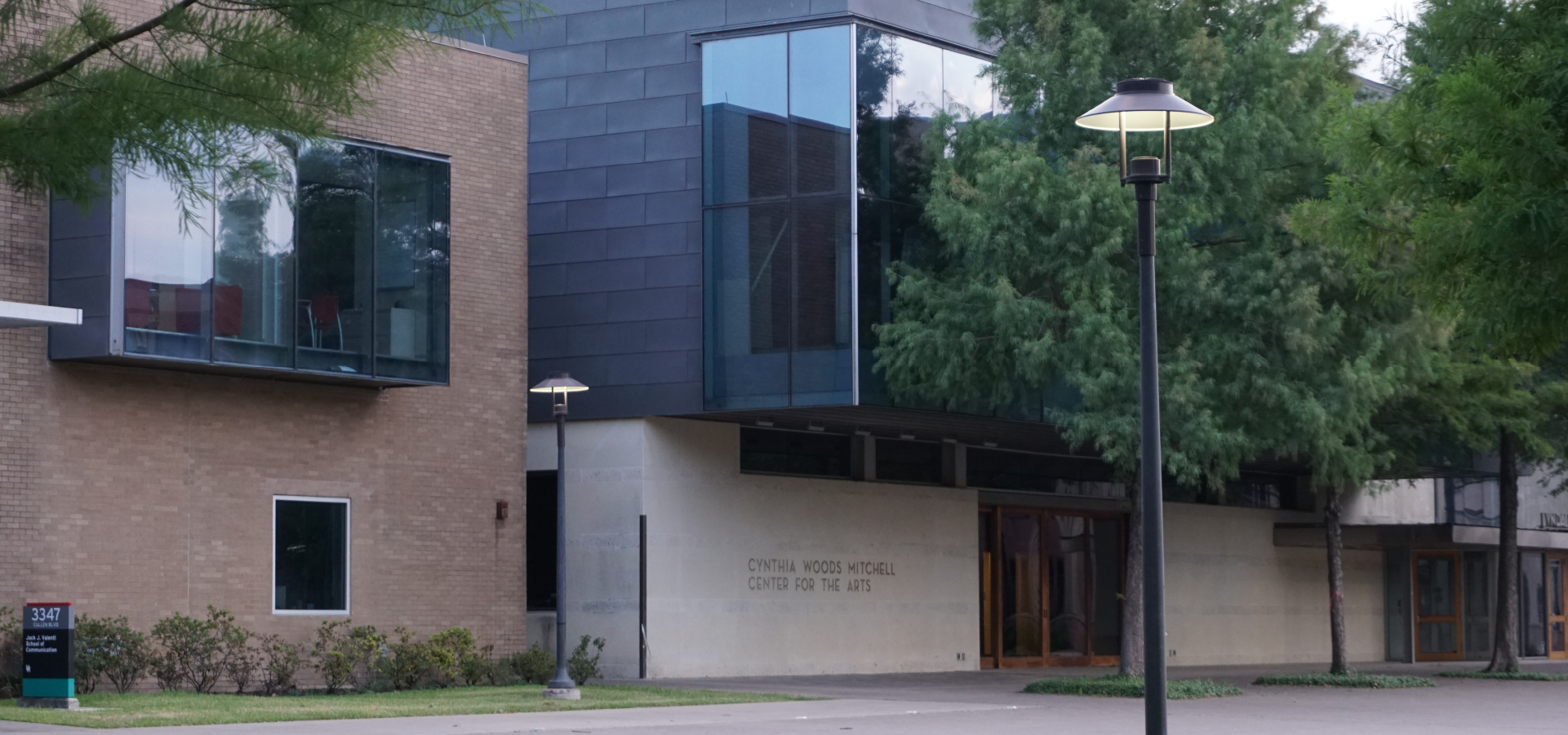 Corner view of the cream and grey façade of the Cynthia Woods Mitchell Center for the Arts Building and a red brick building to its left with trees and a lamppost in the foreground.