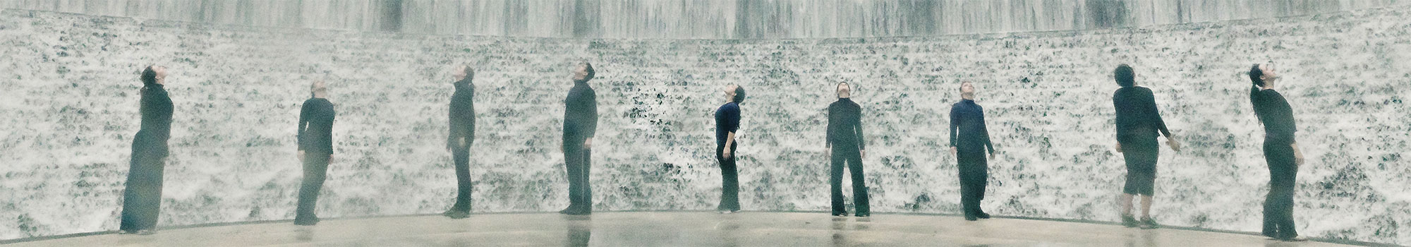 Nine people dressed in black in front of an immense water feature stand spaced apart gazing upward during a performance.