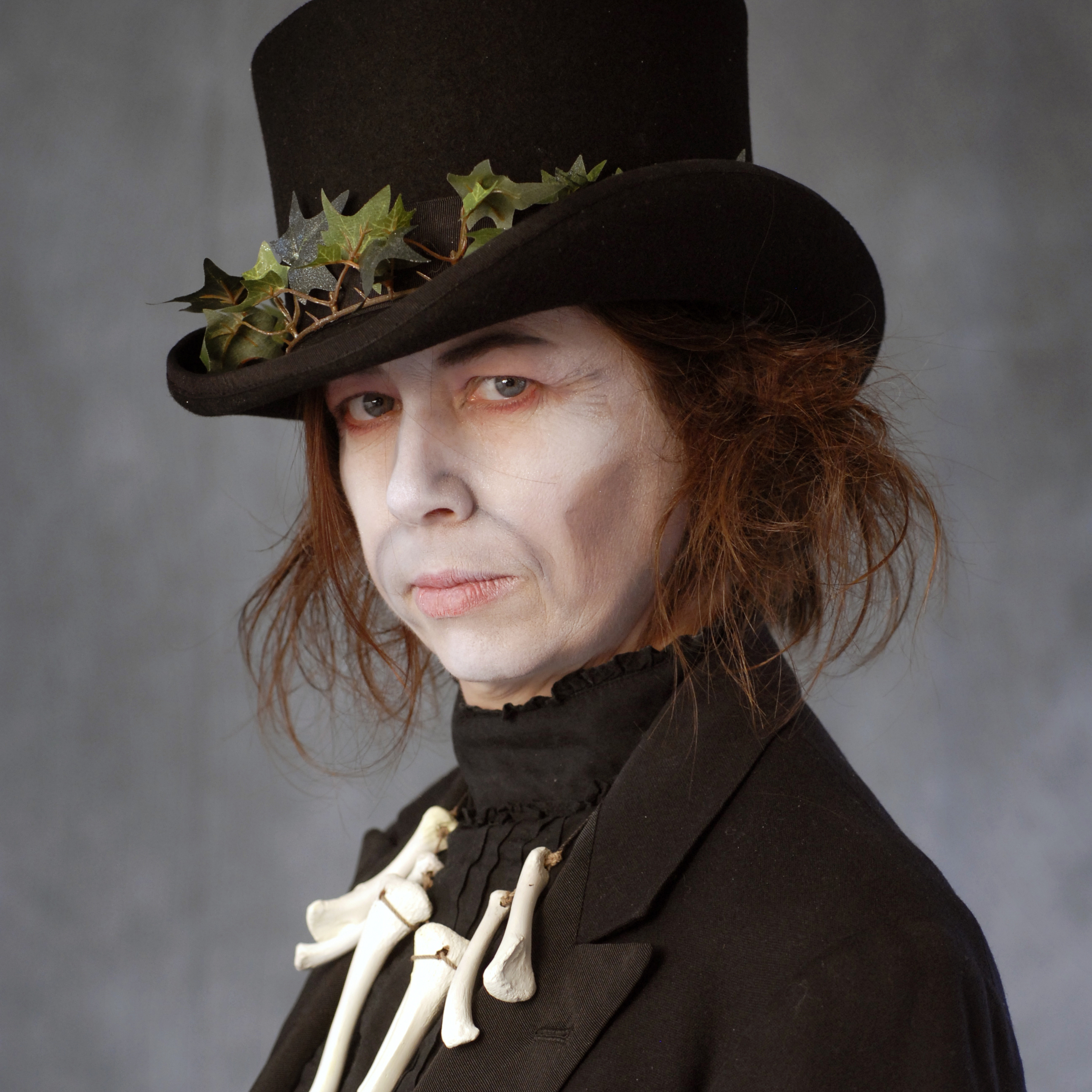 a white woman with chin length brown hair wears a black tophead a necklace that looks like bones. she looks at the camera unsmiling. she has white face makeup on. 