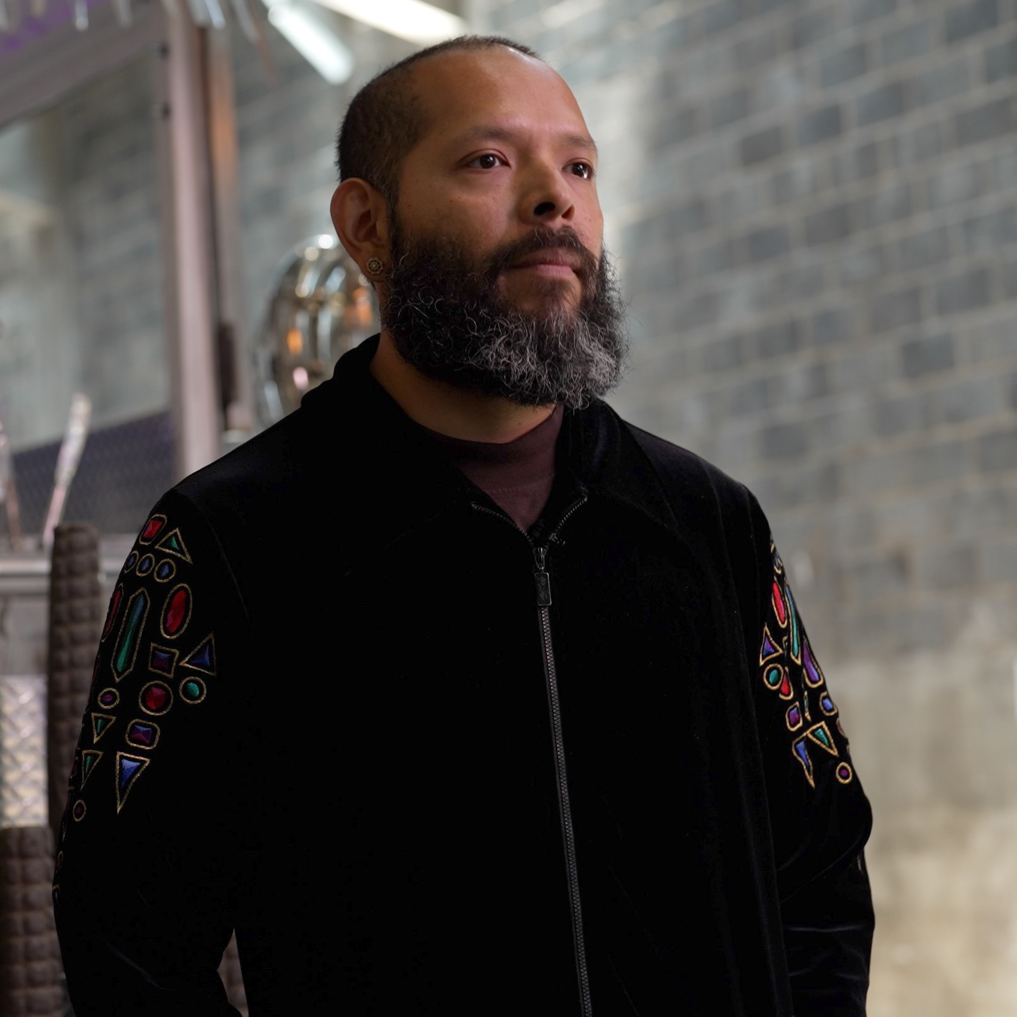 an El Salvadoraian man with a mid-length beard and hair cut close to his head looks away form the camera. he is wearing a black jacket that looks to be made of a reflective material such as satin. there is decorative embroidery or patches near the shoulders. 