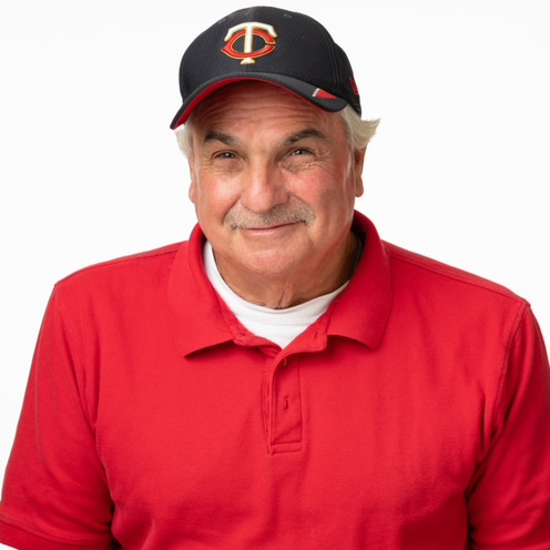  a white man with light gray hair wears a red polo and navy baseball cap. he is posed on a white background and looks into the camera with a soft smile.