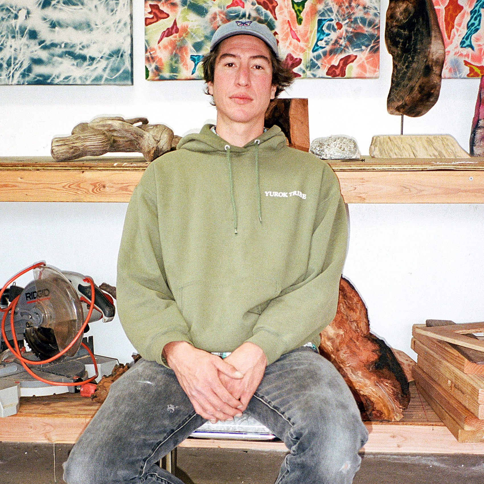 a young man who may be in his early thirties or late twenties has light skin and is wearing jeans and a sage green hoodie. he is sitting on what looks to be a wooden bench inside his art studio.