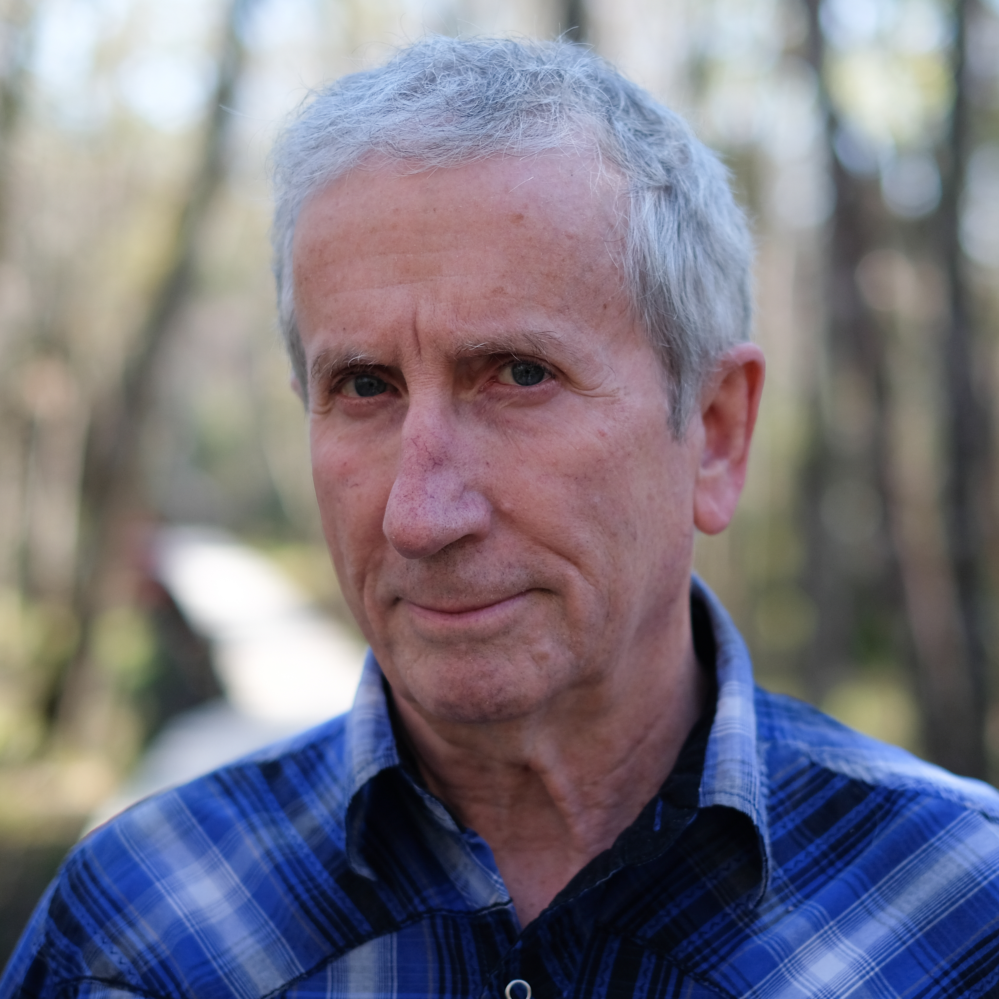  a white man with short light gray hair looks intently into the camera without smiling. he is outside in a wooded area.