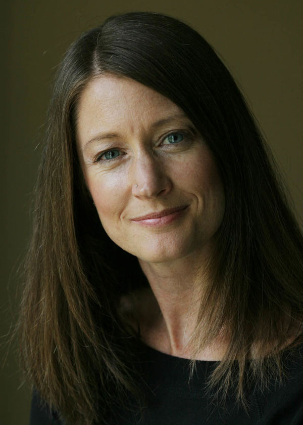 Headshot of a smiling, white-skinned woman with long dark hair wearing a black shirt against a khaki background.