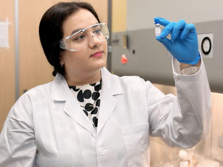 Researcher Ankita Leekha in a lab coat and safety goggles examines a small vial, standing in a laboratory setting.