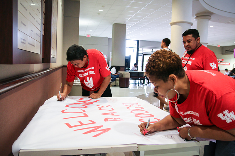 Three people in red shirts stand in front of a large paper banner on a table. Two of the people have markers and are writing on the banner.
