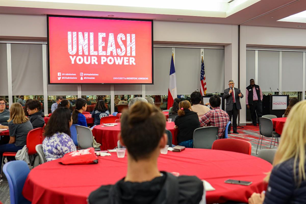 Photograph of a large room filled with people seated at round tables. A man in suit next to screen that says, "Unleash Your Power", are near the back of the room.