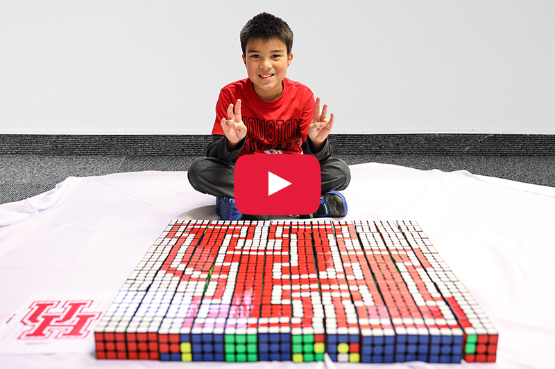 A young Asian boy sits in front of an art piece of the UH logo made of Rubik’s cubes.