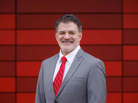 Portrait of a man wearing a grey suit and a red tie smiling while standing in front of a red building.