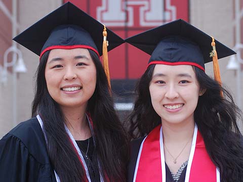 Two Asian women in black graduation regalia smile together. A large red UH banner is on the building behind them.