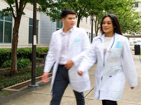 Male and female students wearing lab coats walking past a building. A motion blur effect is applied to the students.